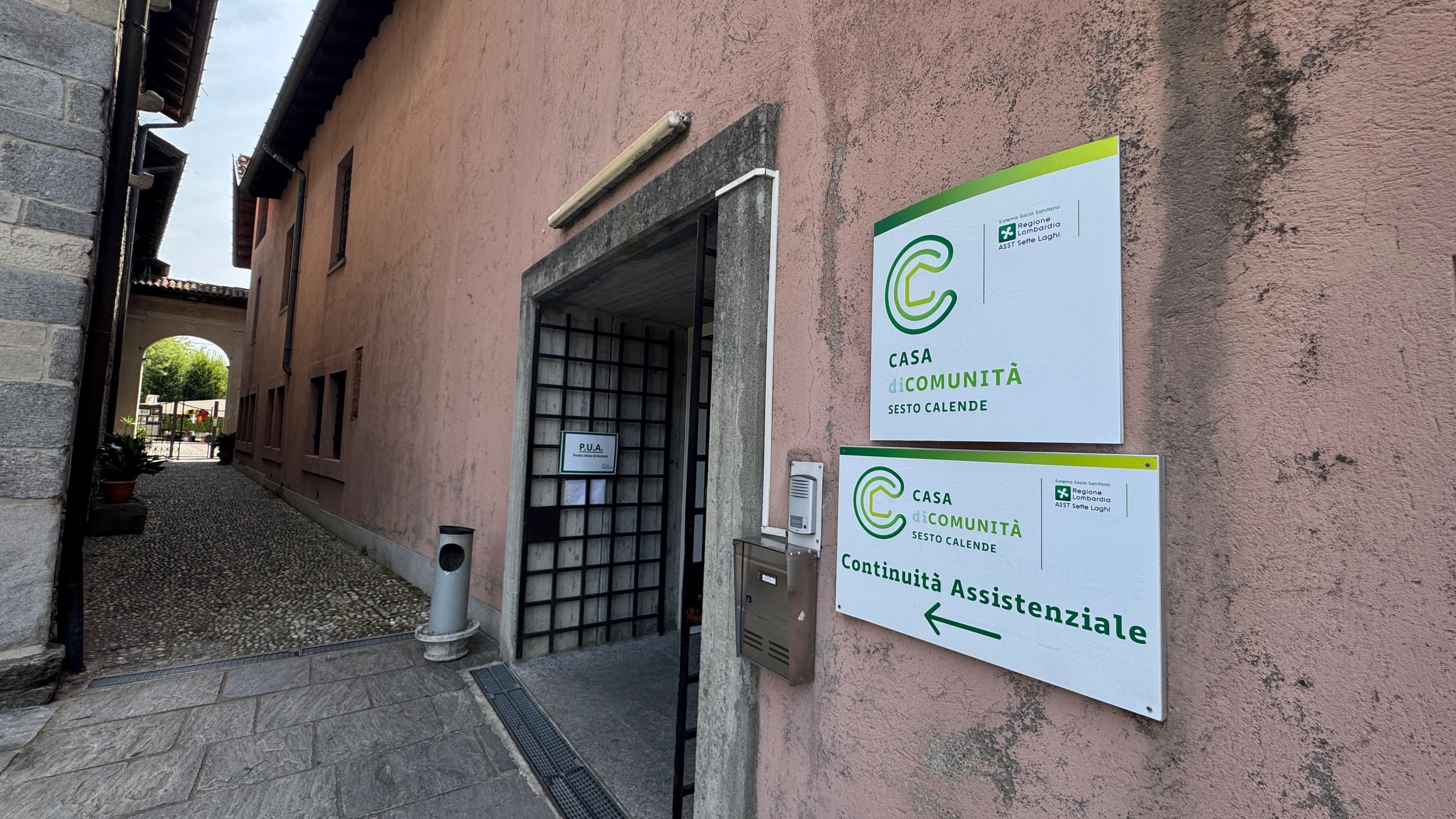 Vista dell’ingresso della Casa di Comunità di Sesto Calende. Sulla parete dell’edificio sono presenti le insegne della struttura sanitaria e della Continuità Assistenziale, con accesso da un passaggio pedonale lastricato