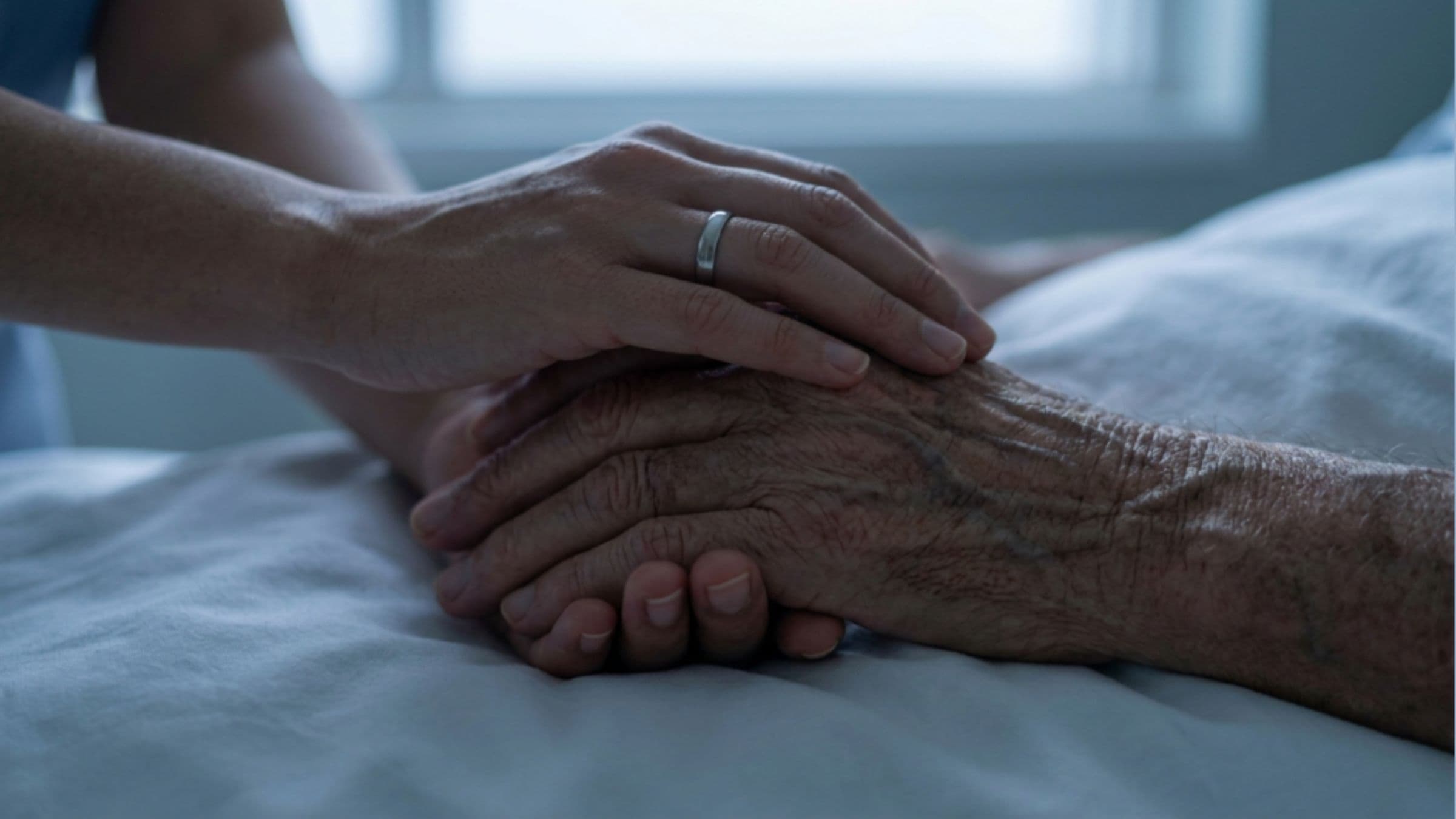 Mani di una persona che stringono e accarezzano la mano di una persona anziana, in un gesto di vicinanza e assistenza, immagine rappresentativa del tema della morte medicalmente assistita.