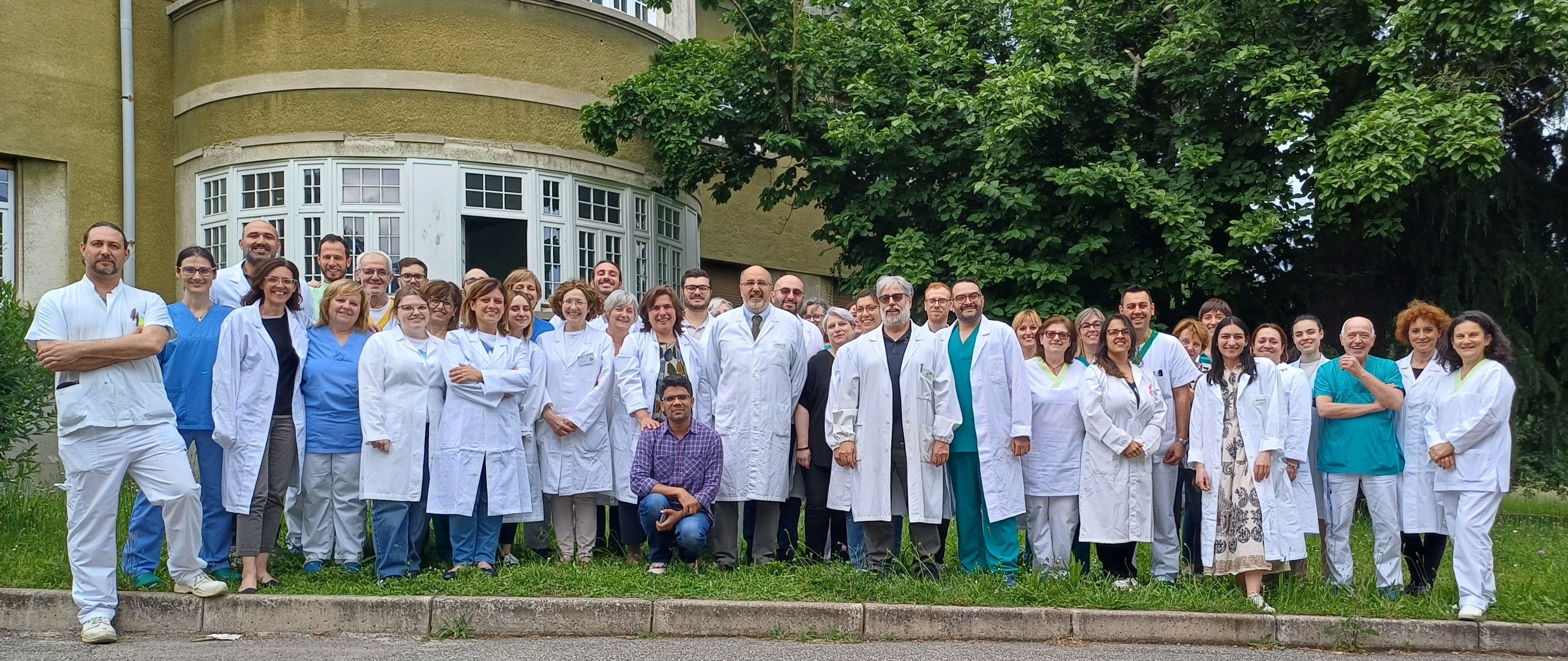 Foto di gruppo dell’équipe di Anatomia Patologica, composta da numerosi professionisti sanitari in camice bianco e divisa, riuniti all’aperto davanti a un edificio ospedaliero circondato dal verde.